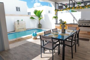A poolside dining area with a glass table, chairs, and a grill.