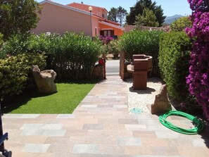Ein gepflasterter Weg führt zu einem Gebäude mit einer Steinmauer und einem Garten mit Sträuchern und Blumen.