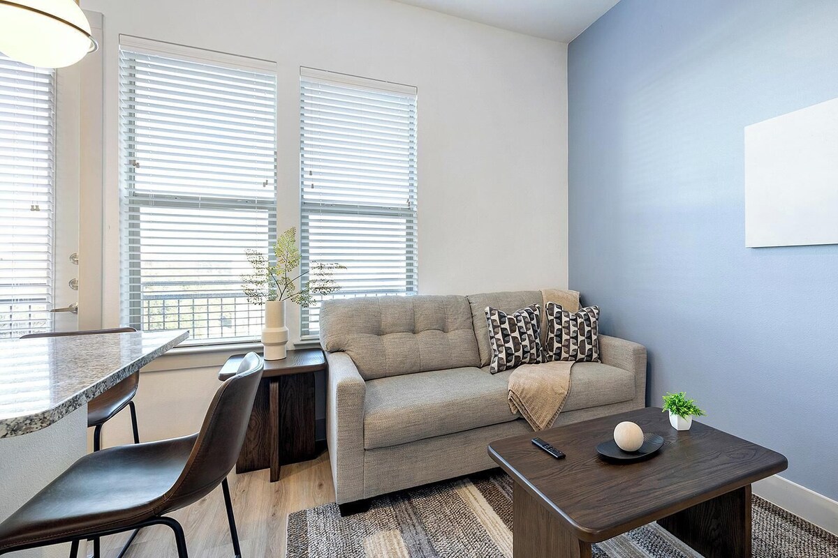 Ein Wohnzimmer mit einer Couch, einem Couchtisch, einem Essbereich mit Stühlen und einem großen Fenster mit Jalousien.