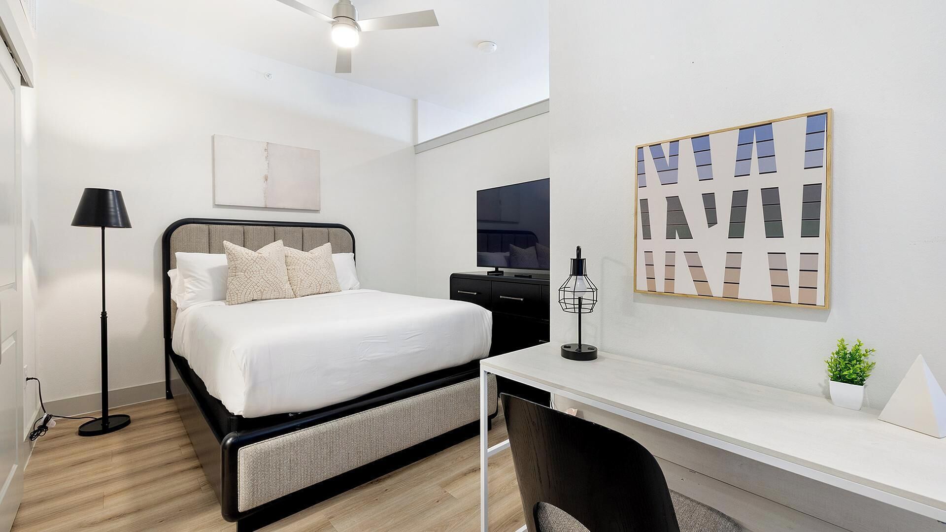 A modern hotel room with a bed, desk, and chair, a ceiling fan, and a framed artwork on the wall.