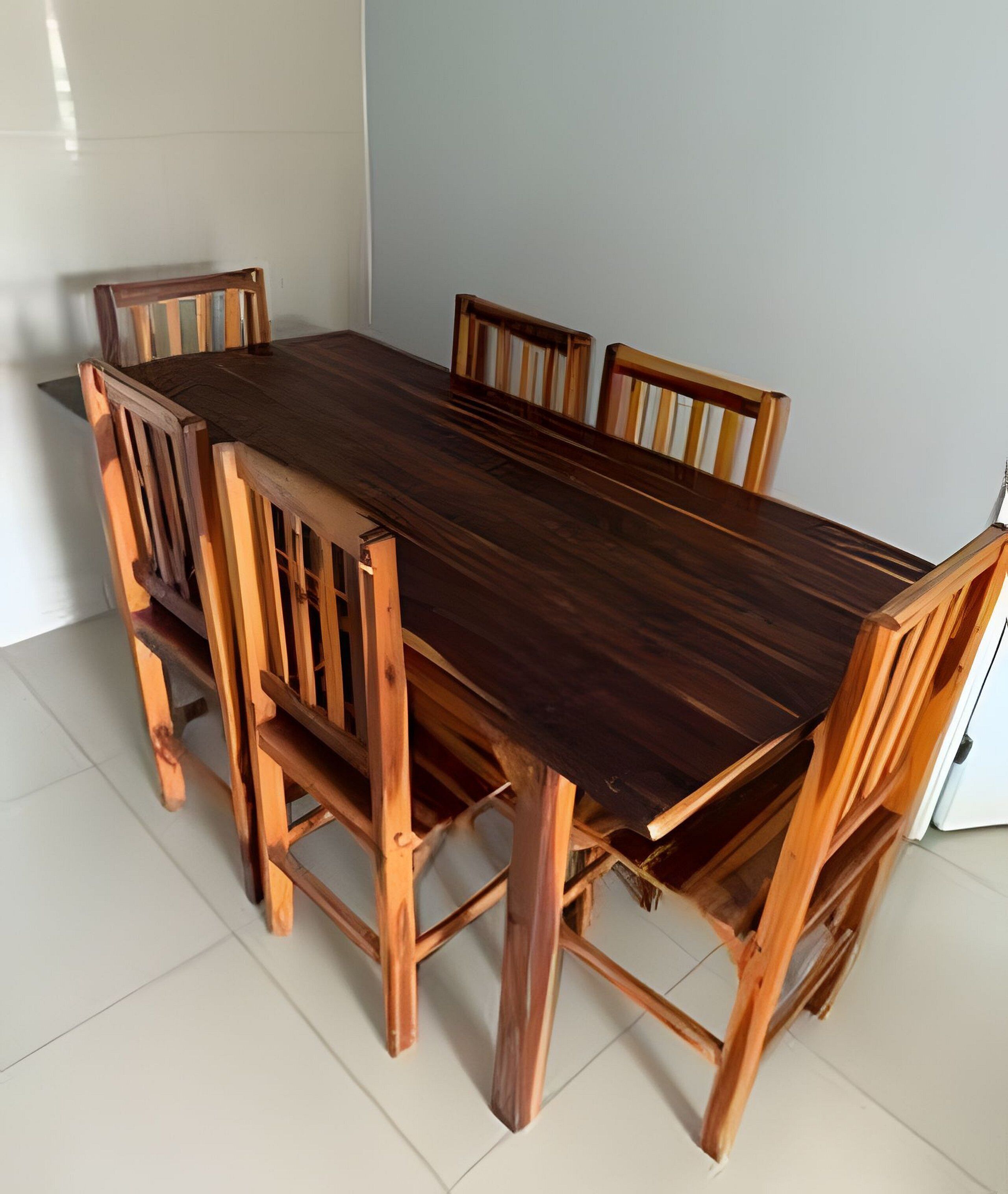 Una mesa rectangular de madera con seis sillas, ubicada en una habitación con piso de baldosas blancas y paredes de color claro.