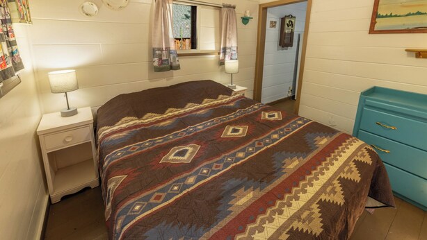 Une chambre à coucher comprenant un lit, une table de chevet, une lampe, une commode et une fenêtre avec des rideaux.