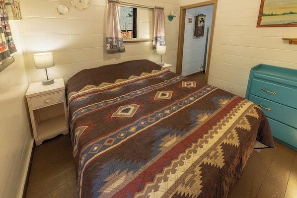 A bedroom with a bed, a nightstand, a lamp, a dresser, and a window with curtains.