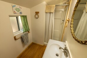 A bathroom with a wooden floor, a white sink with a gold faucet, a gold-framed mirror, a shower with a white curtain, and a window with a green curtain.