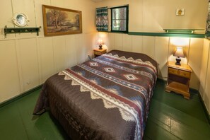 Un dormitorio con una cama, dos mesitas de noche, un velador, un cuadro, una ventana con cortinas y un espejo montado en la pared.