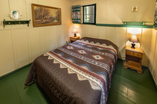 A bedroom with a bed, two bedside tables, a nightstand, a painting, a window with curtains, and a wall-mounted mirror.