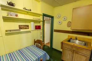 Una habitación pequeña y acogedora con una cama individual, una silla de madera, una mesita, un lavamanos y un armario. Se ven estantes con diversos objetos y una puerta que da a otra habitación.