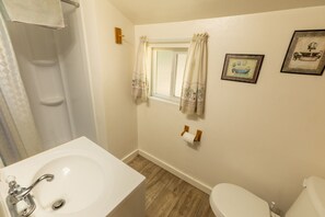 A bathroom with a sink, toilet, shower, and a window with curtains.