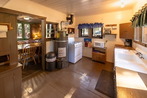 A compact kitchen with a stove, microwave, sink, and a dining area with chairs. - Two-Story Pine Cabin at Camp Layman | Spacious Mountain Lodge for Eight (Blairsden-Graeagle)