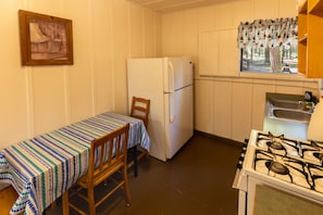 A compact kitchen with a refrigerator, a gas stove, a window with a view, and a table with a striped tablecloth. - Accessible Cub Cabin at Camp Layman | Playful & Inclusive Riverside Stay (Blairsden-Graeagle)