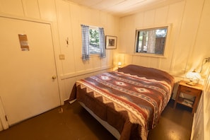 A single bed with a patterned bedspread, a window with a view of trees, a bedside lamp, and a door with a small plaque. - Accessible Cub Cabin at Camp Layman | Playful & Inclusive Riverside Stay (Blairsden-Graeagle)