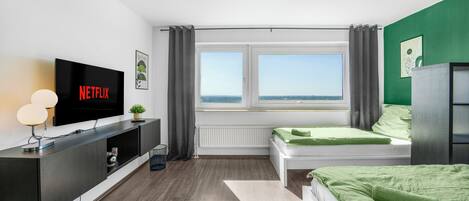 Un dormitorio moderno con un televisor que muestra 'NETFLIX', una cama con ropa de cama verde, un ventanal con vistas y una planta pequeña en la mesita de noche.