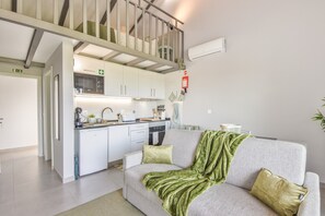 Eine moderne Küche mit weißen Schränken, einer Mikrowelle und einem Spülbecken. Ein graues Sofa mit einer grünen Tagesdecke und zwei Kissen. Im Hintergrund ist eine Treppe mit Geländer zu sehen.