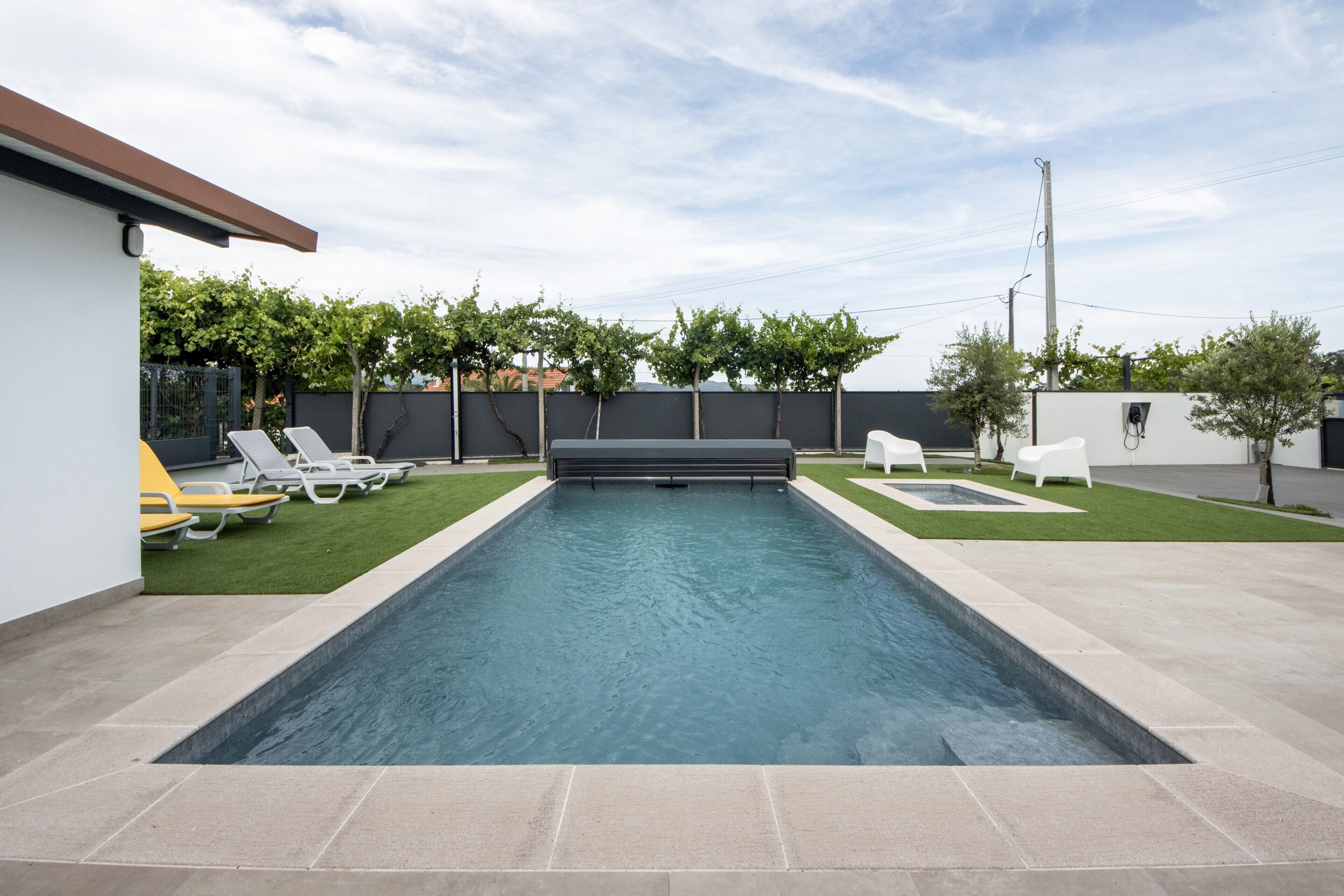 Une piscine rectangulaire à bordure sombre, entourée d’une terrasse carrelée. On y trouve des chaises longues et un petit espace herbeux agrémenté d’arbres.