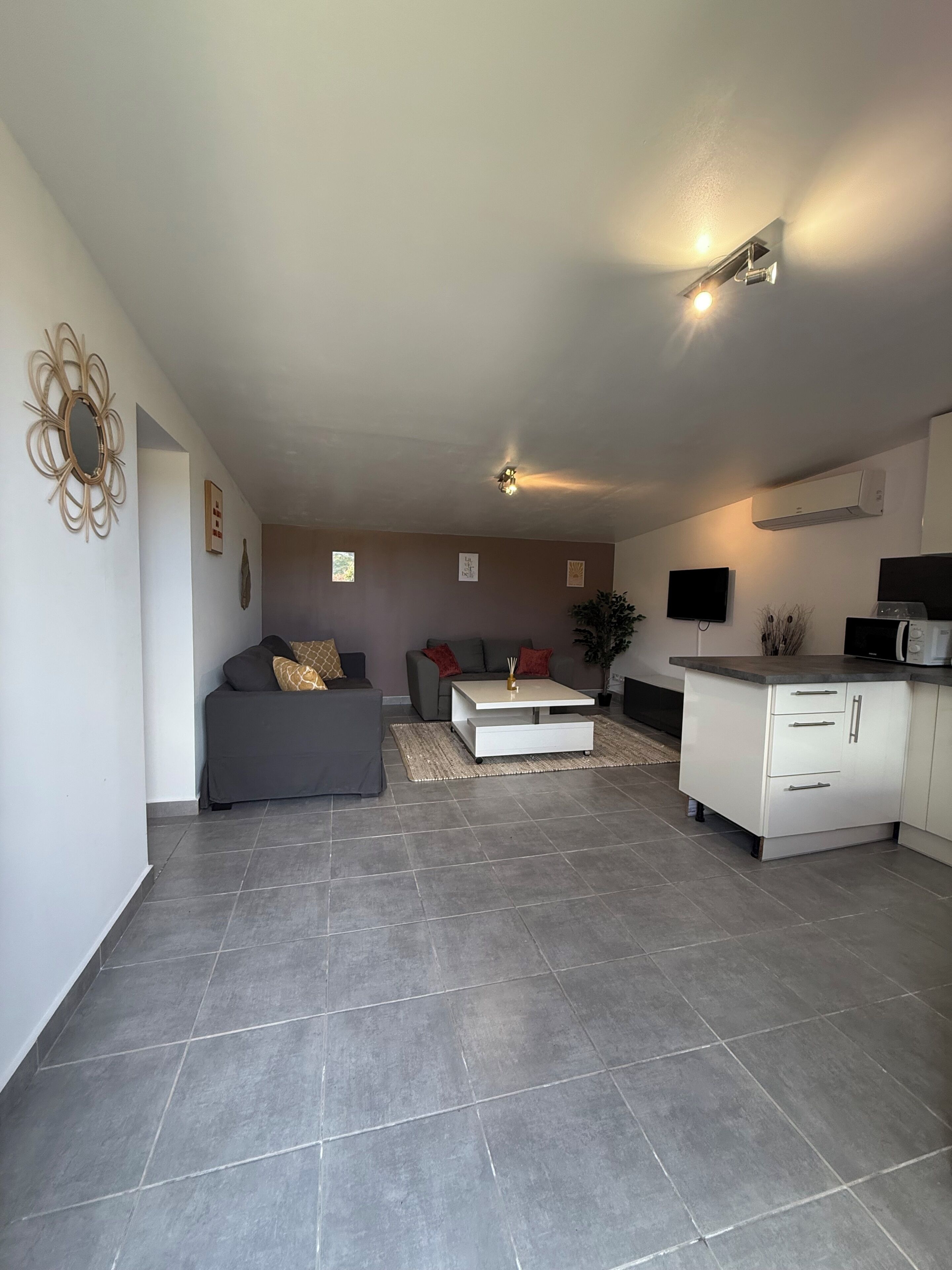 Una sala de estar moderna con un sofá gris, una mesa de centro blanca y una cocina con un armario blanco y microondas.