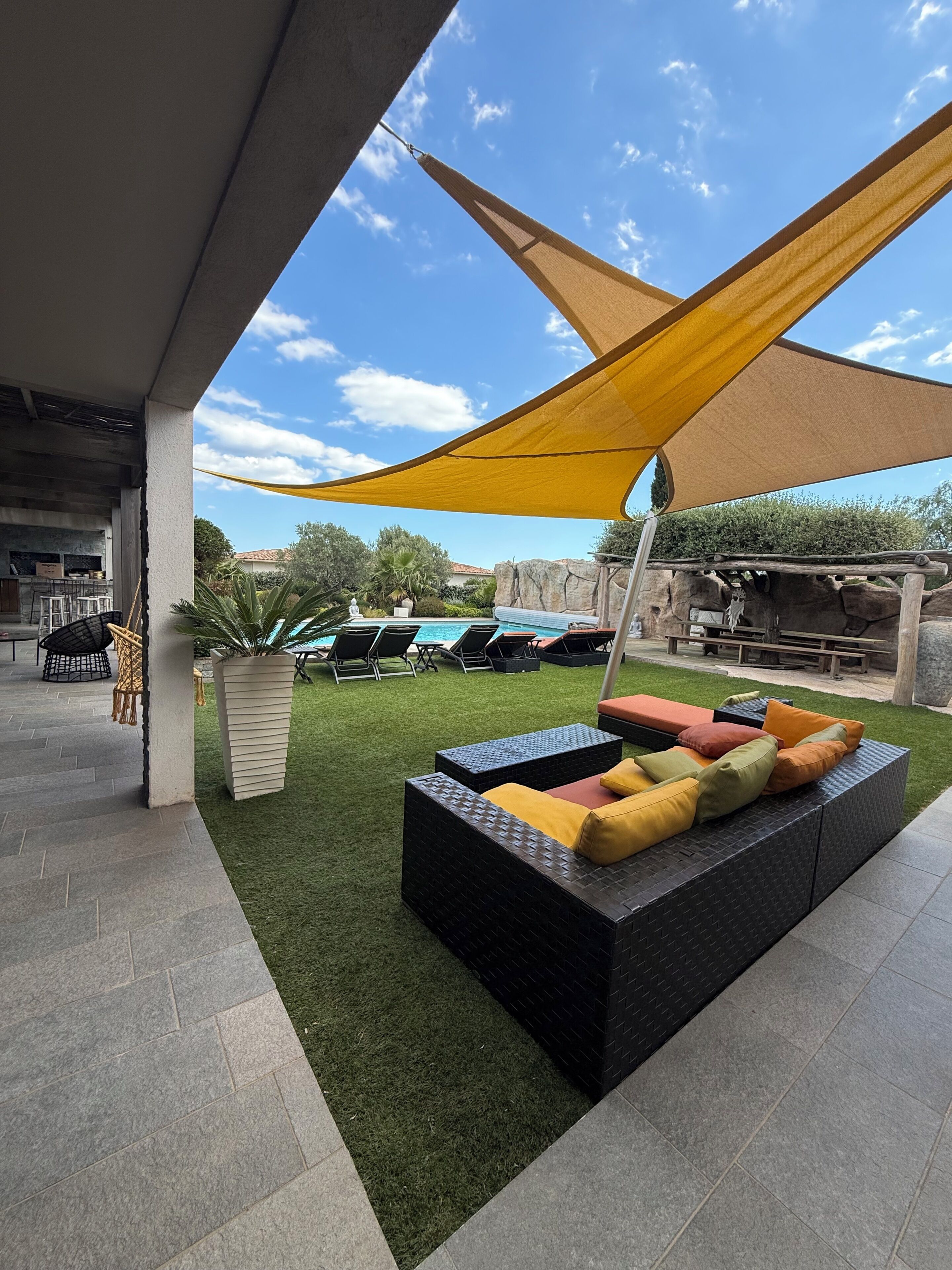 Un espacio al aire libre con sillones y cojines, una piscina y una zona sombreada con un toldo amarillo.