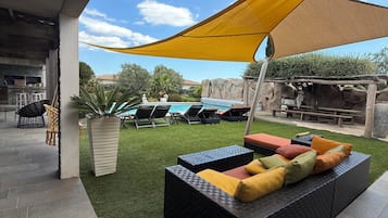Un espacio al aire libre con sillones y cojines, una piscina y una zona sombreada con un toldo amarillo.