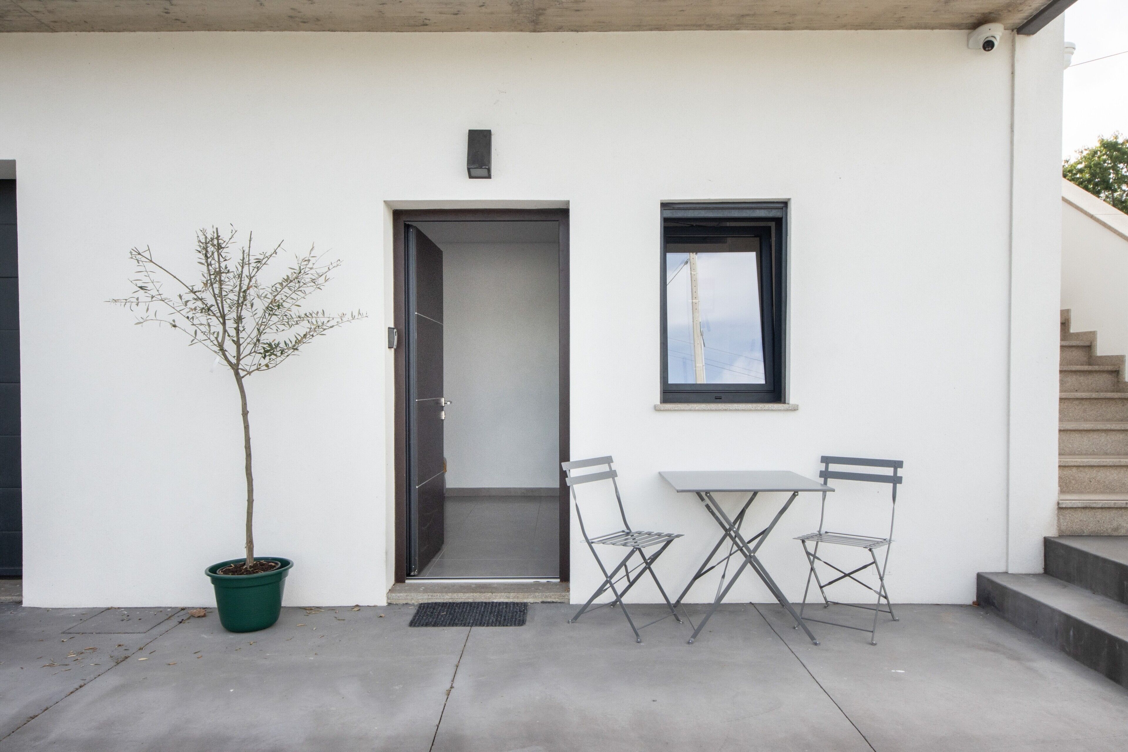 A white building with a door, a window, a potted tree, and a table with chairs.