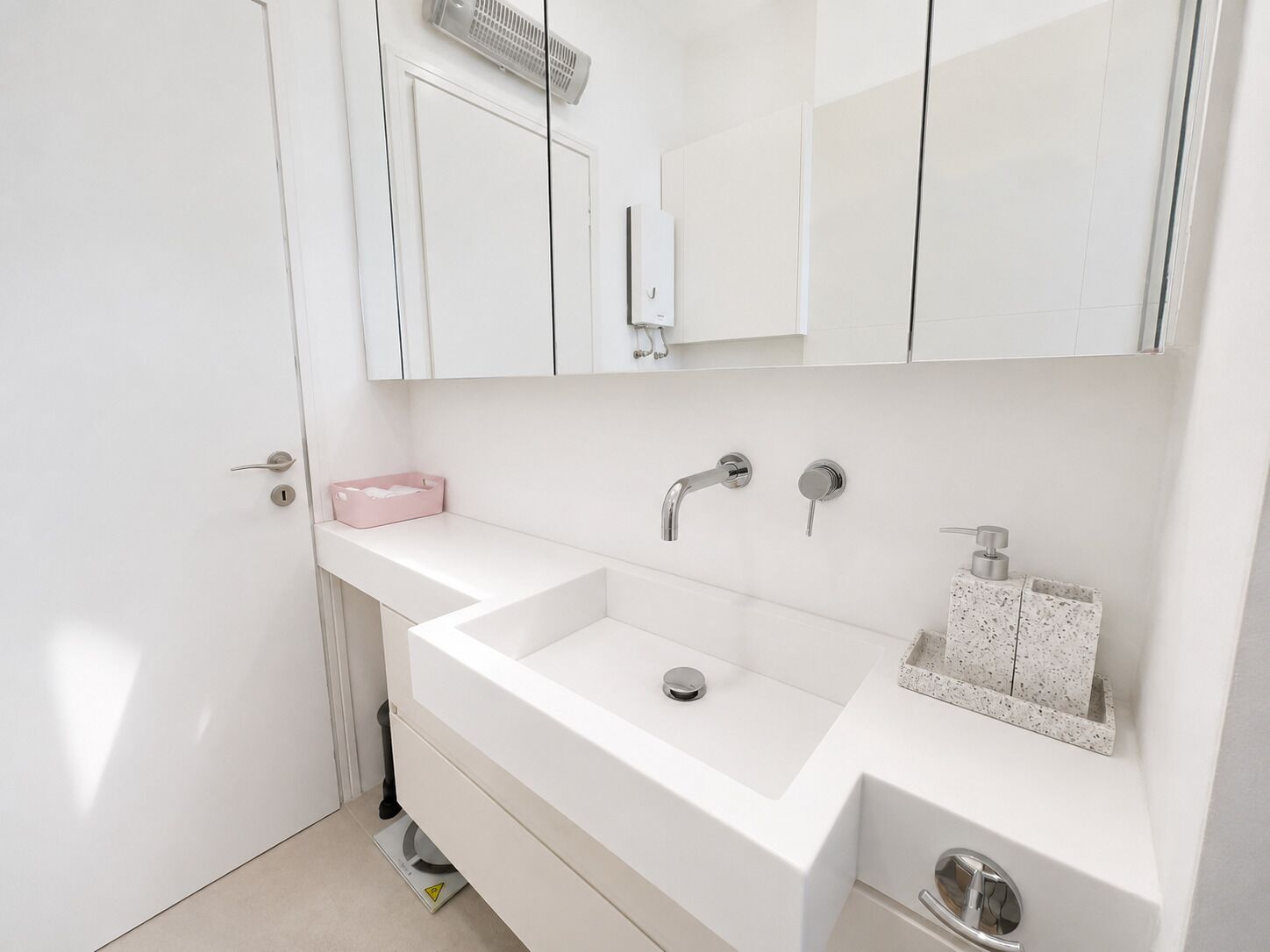 Een moderne badkamer met een witte wastafel, twee kranen, een spiegel en een klein roze zeepbakje.