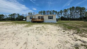A single-story house with a front deck, surrounded by a sandy area and trees. - 2-bedroom mobile home a minute off the Suncoast Parkway. Comfy and cozy! (Homosassa)
