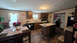 A compact kitchen with wooden cabinets, a dining table with chairs, a central island, and a refrigerator. - 2-bedroom mobile home a minute off the Suncoast Parkway. Comfy and cozy! (Homosassa)