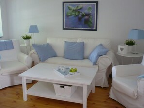 Ein helles Wohnzimmer mit einem weißen Sofa, zwei blaue Karokissen, einem weißen Couchtisch mit einer Obstschale und einem gerahmten Bild an der Wand.