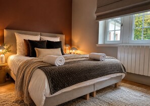 Un lit bien fait, avec une couverture matelassée, des oreillers et des serviettes enroulées. On y trouve une table de chevet avec une lampe, une fenêtre donnant sur la verdure et un radiateur.