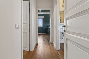 Un couloir avec des portes blanches, un plancher en bois et une vue sur une pièce dont un mur est bleu et qui possède une fenêtre.