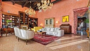 Una habitación amplia con un sofá blanco, un tapete rojo, una mesa de centro de madera, un sillón blanco, una estantería, un armario y una araña.