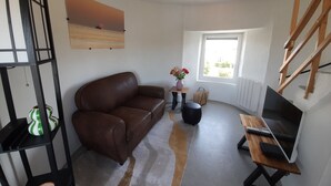 Un salon avec un canapé en cuir marron, une petite table sur laquelle est posé un vase de fleurs, un téléviseur sur un meuble en bois et une fenêtre donnant sur un paysage verdoyant.
