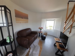 Un salon avec un canapé en cuir marron, une petite table sur laquelle est posé un vase de fleurs, un téléviseur sur un meuble en bois et une fenêtre donnant sur un paysage verdoyant.