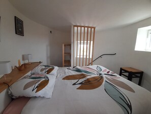 A bedroom with a bed, a wooden side table, a desk with a lamp, a window, and a door.