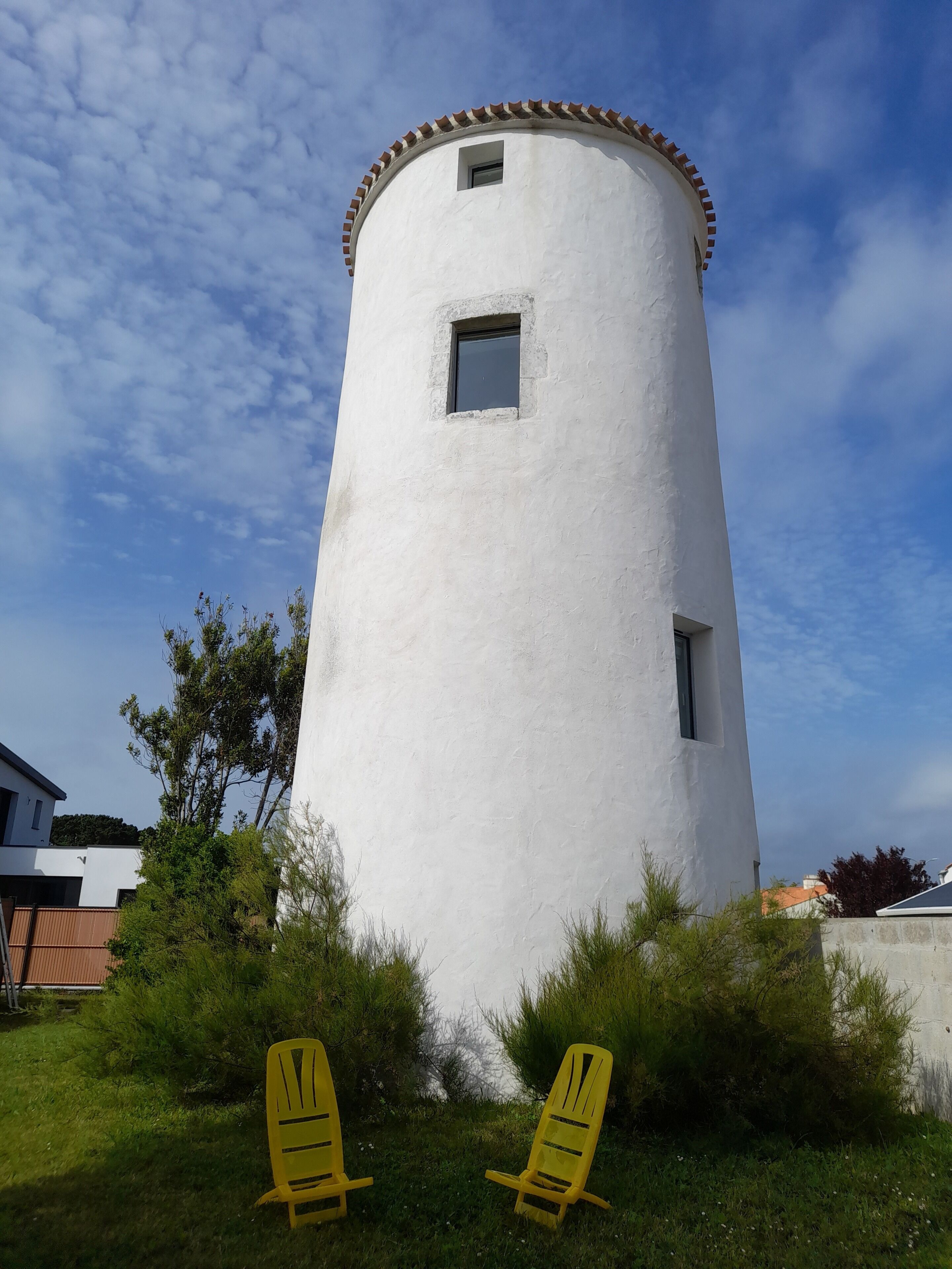 A white cylindrical tower with two windows, surrounded by greenery and yellow chairs.