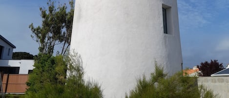 A white cylindrical tower with two windows, surrounded by greenery and yellow chairs.