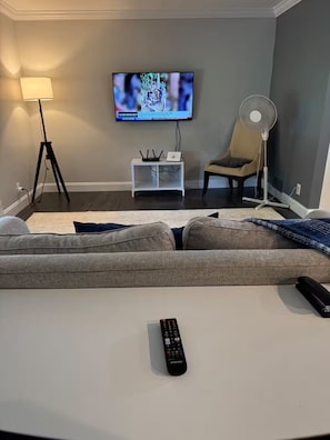 Ein Wohnzimmer mit einer Couch, einem Fernseher, einer Lampe, einem Ventilator und einer Fernbedienung.