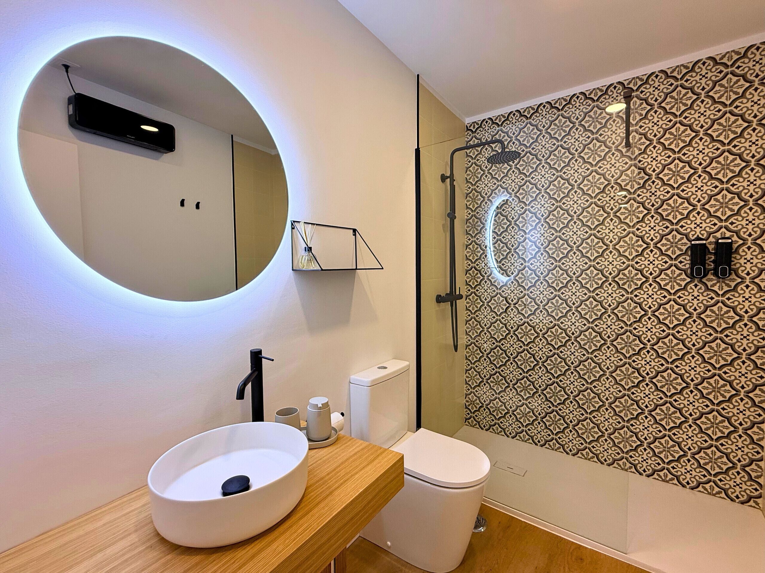 Ein modernes Badezimmer mit rundem Spiegel, einem Aufsatzwaschbecken, einer Toilette, einer Duschkabine und Wandfliesen mit Muster.