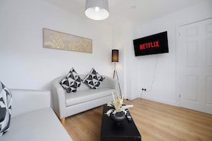 Ein modernes Wohnzimmer mit einem weißen Sofa, geometrisch gemusterten Kissen, einem schwarzen Couchtisch mit einer Vase, einer Stehlampe und einem angebrachten Fernseher, der 'NETFLIX' zeigt.