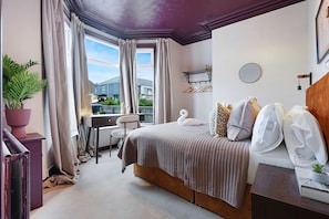 Quarto com cama, escrivaninha, cadeira, janela com cortinas, planta em vaso e vista de um edifício.