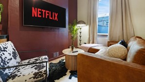 Un soggiorno con una TV che mostra 'NETFLIX', un divano, una poltrona, una lampada e una finestra con tende.