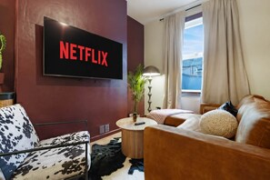 Uma sala de estar com uma TV exibindo 'NETFLIX', um sofá, uma poltrona, um abajur e uma janela com cortinas.
