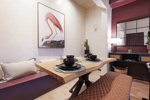 Uma área de refeições com uma mesa de madeira posta para dois, um banco acolchoado, uma planta de cacto e uma obra de arte na parede com um flamingo.