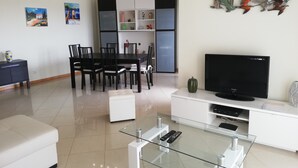 Uma sala moderna com uma mesa de centro de vidro, uma televisão de tela plana, uma área de jantar com cadeiras e um sofá branco.