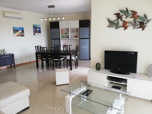 Ein modernes Wohnzimmer mit einem Glastisch, einem Flachbildfernseher, einem Essbereich mit Stühlen und einem weißen Sofa.