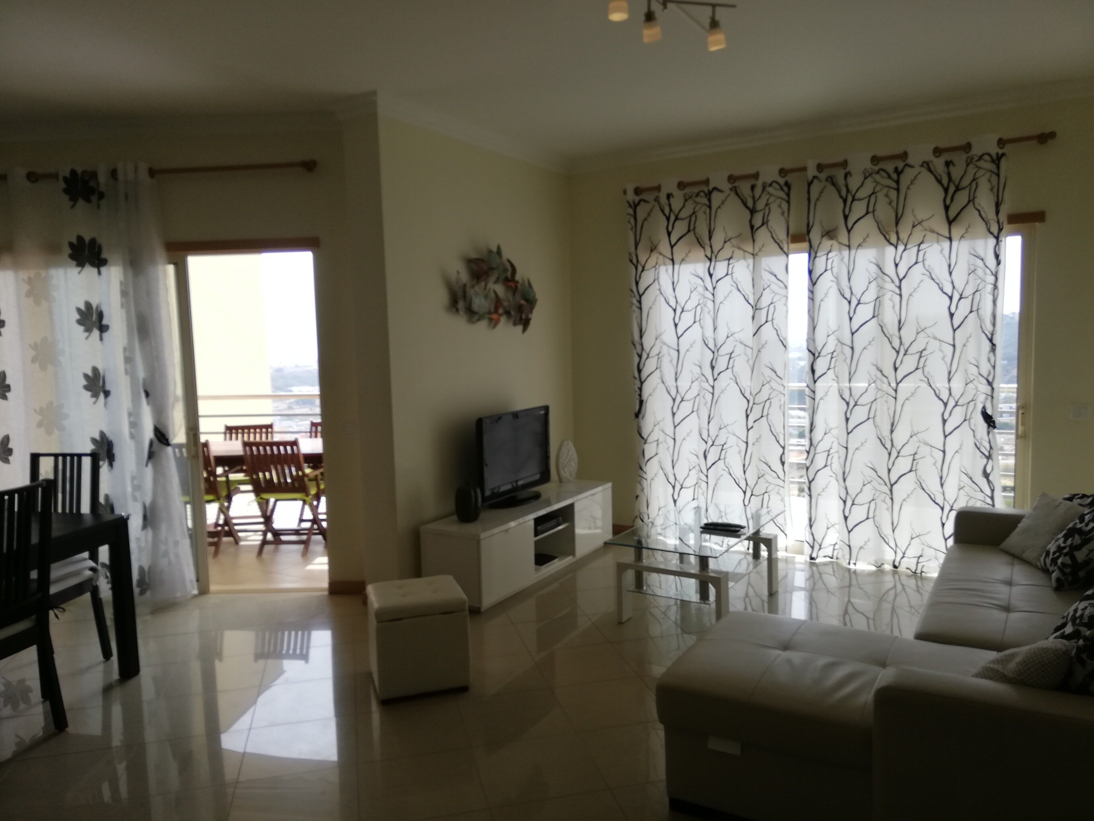 Un salón con sofá, televisión, mesa de centro de vidrio, comedor con sillas y vistas al exterior.