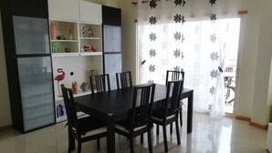 Uma área de refeições com mesa e cadeiras, um armário branco com prateleiras e uma janela com cortina florida.