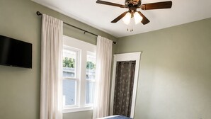 Un dormitorio con cama, ventilador de techo, televisor y ventana con cortinas.