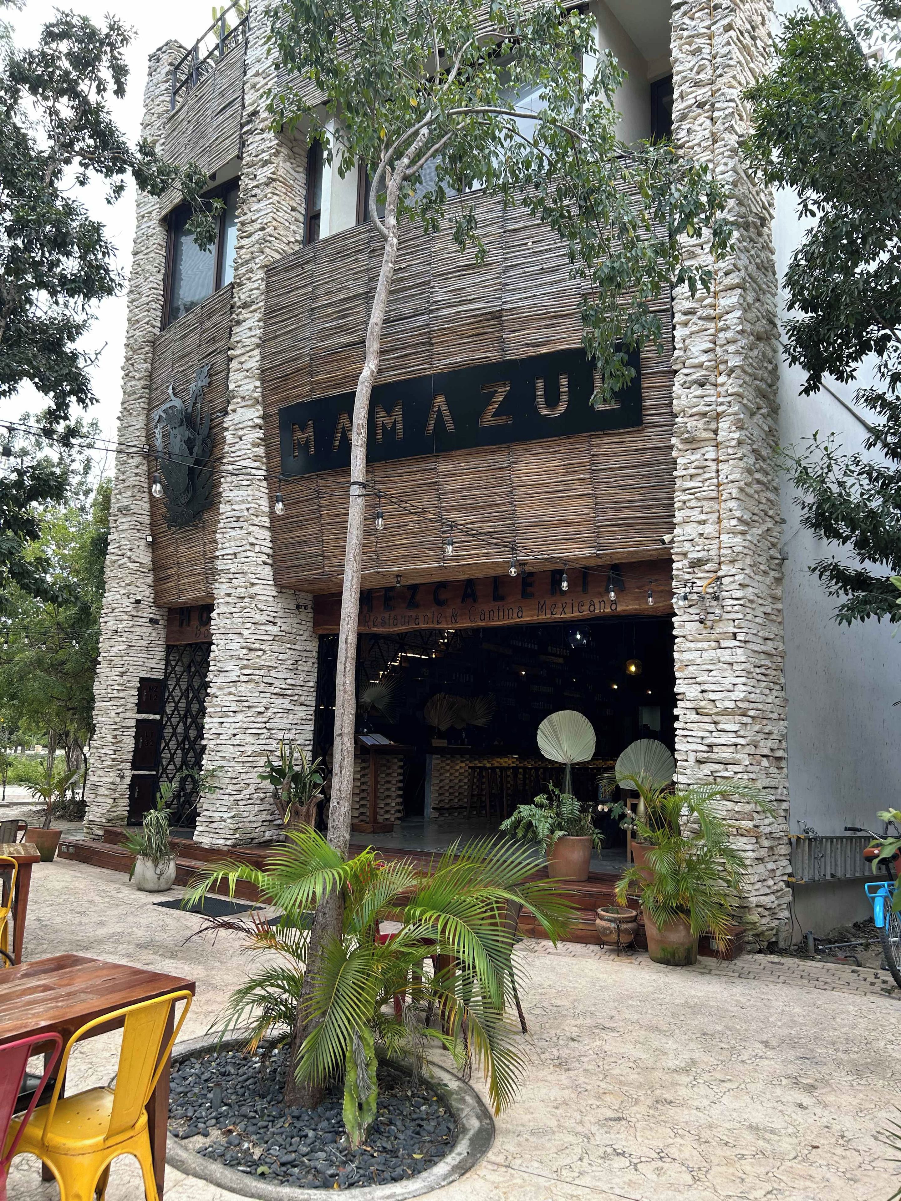 A building with a stone facade, a sign reading 'MAMA ZU', and outdoor seating with potted plants.