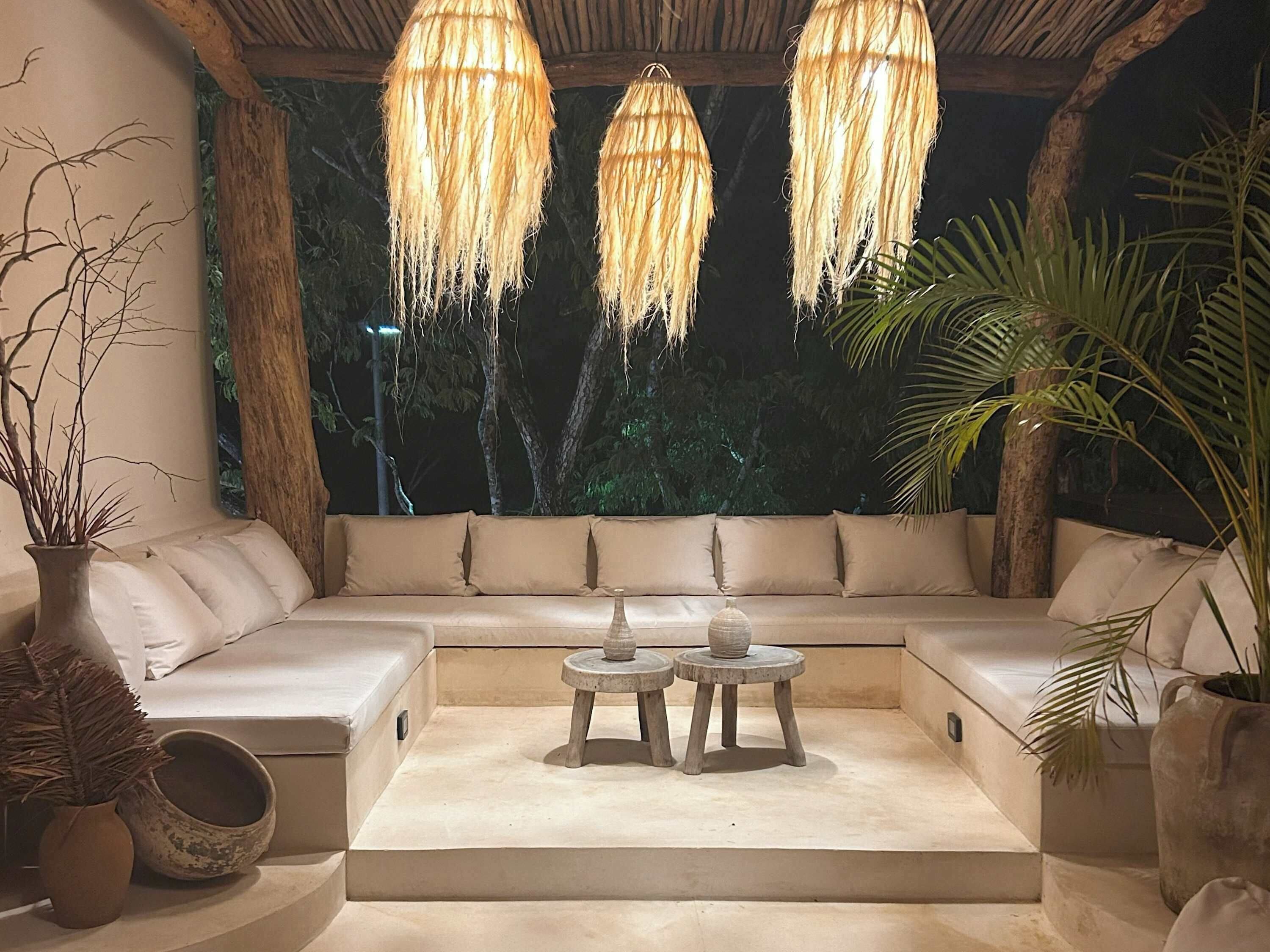 A cozy outdoor seating area with a cushioned sofa, two small round tables, and hanging wicker lamps.