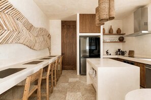 A modern kitchen with a central island, wooden chairs, a stainless steel refrigerator, and a hanging light fixture. - Tulumaya Boho Luxe Villa in Aldea Zama Concierge (Tulum)