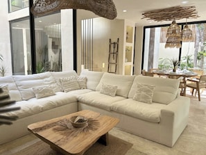 A modern living room with a sectional sofa, a wooden coffee table, and a dining area with a table set for four. - Tulumaya Boho Luxe Villa in Aldea Zama Concierge (Tulum)